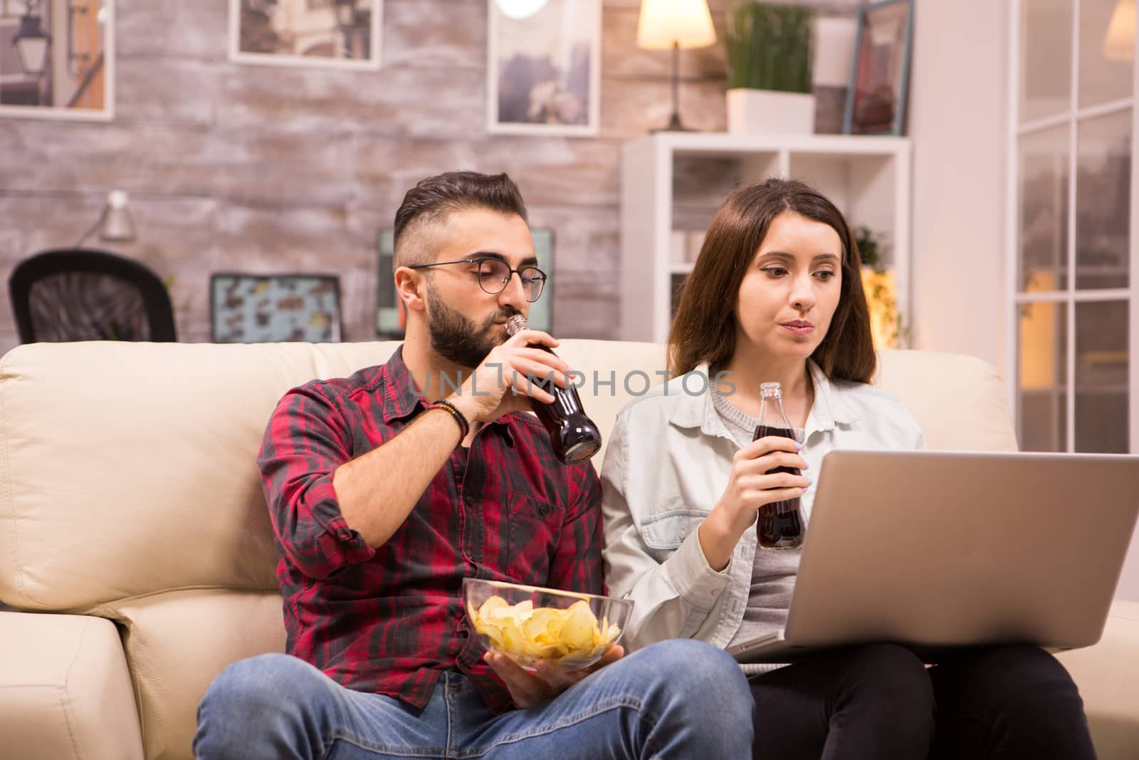 Couple watching a movie sitting comfortable on couch eating chip and drinking soda.