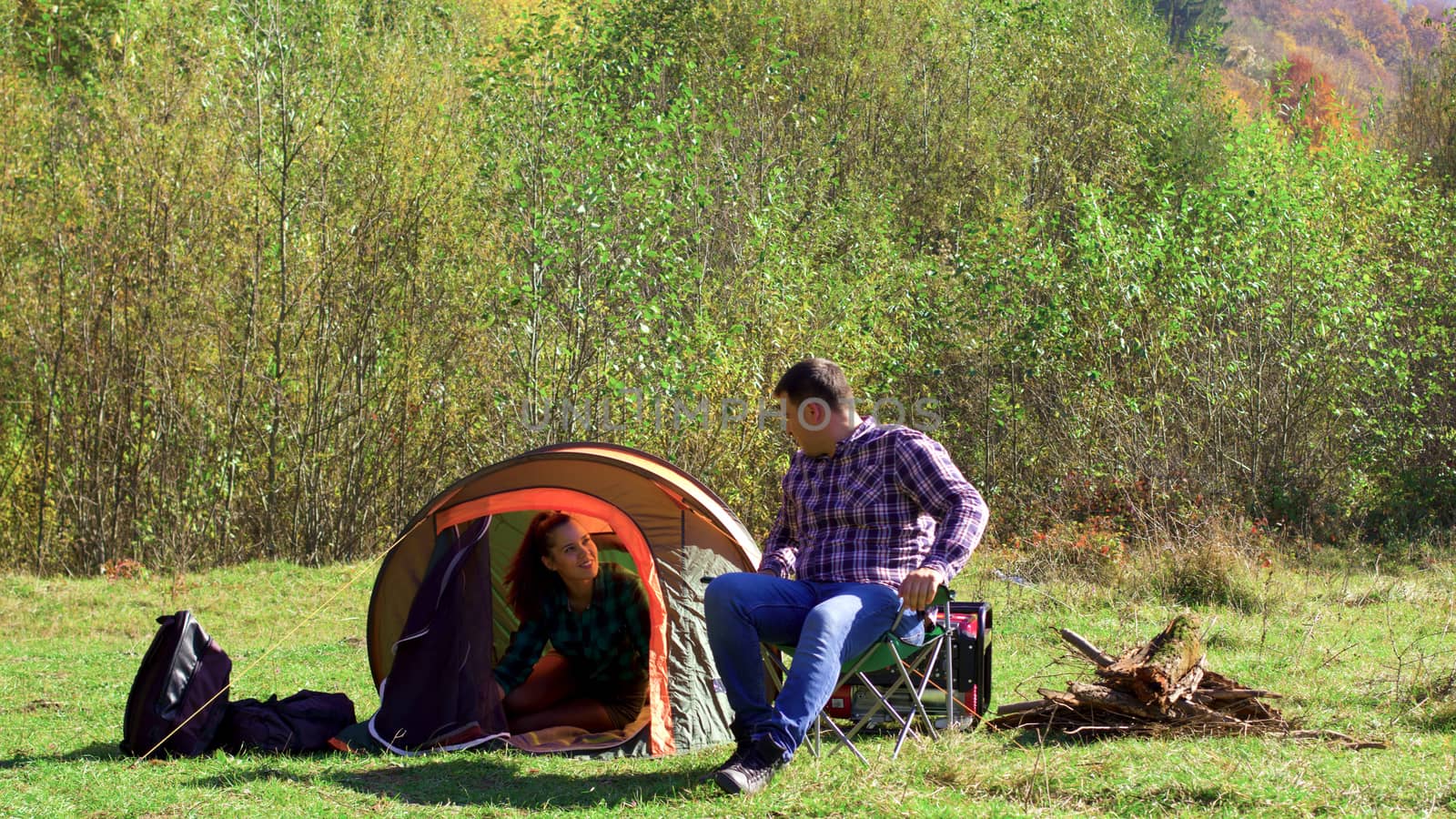 Boyfriend sitting on camping seat while girlfriend preparing the tent for camping. Young couple in the wood.