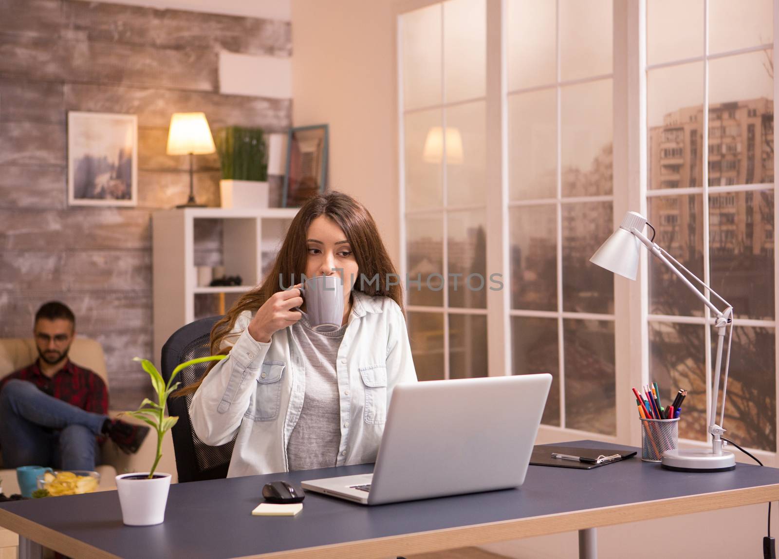 Female entrepreneur working on laptop from home and drinking coffee. Boyfriend relaxing on sofa in the background.