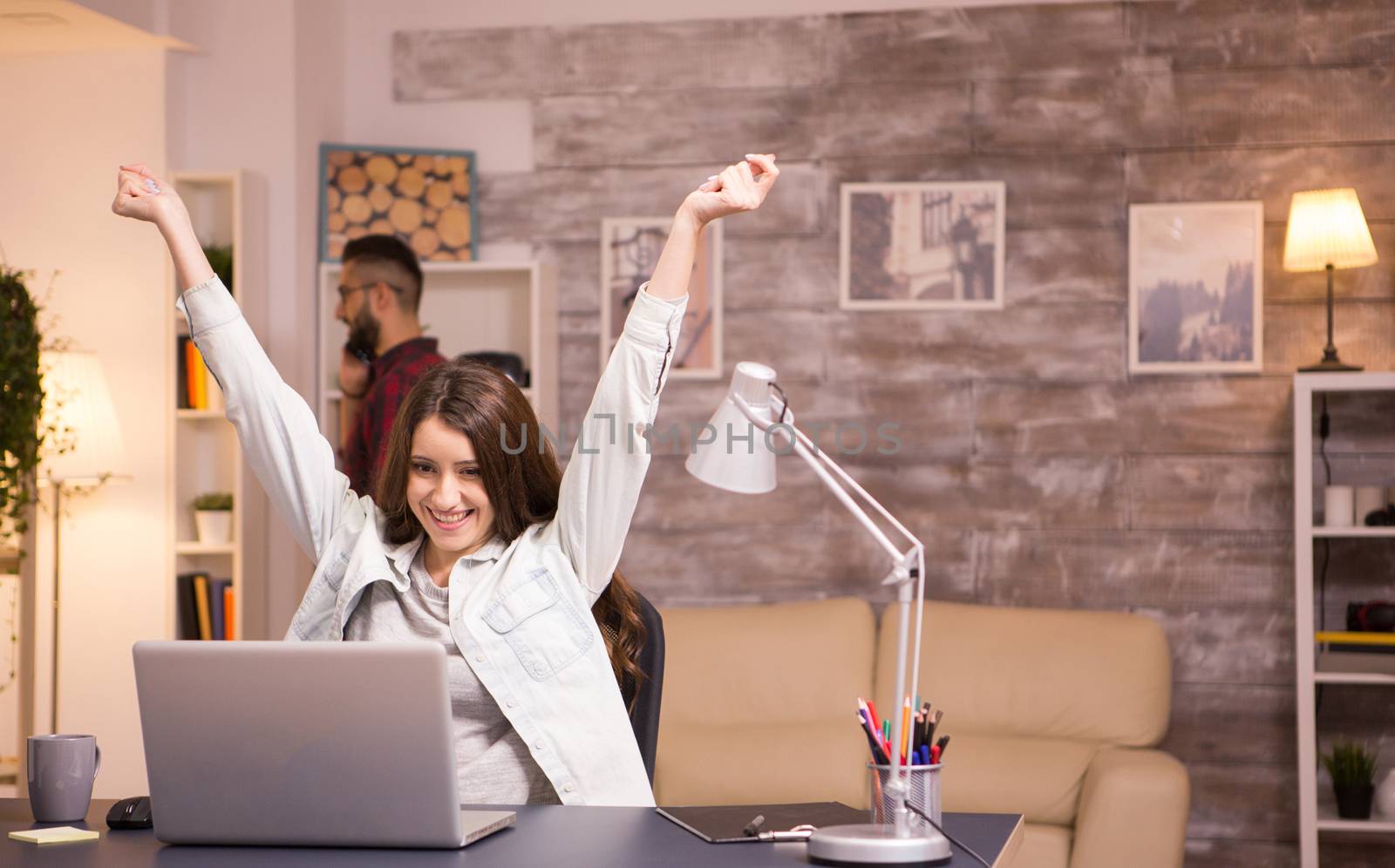 Happy young woman with hands raised while working on laptop in living room. Boyfriend talking on the phone in the background.