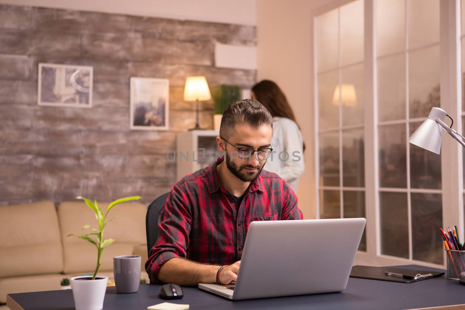 Bearded freelancer working typing on laptop from home. Girlfriend in the background.