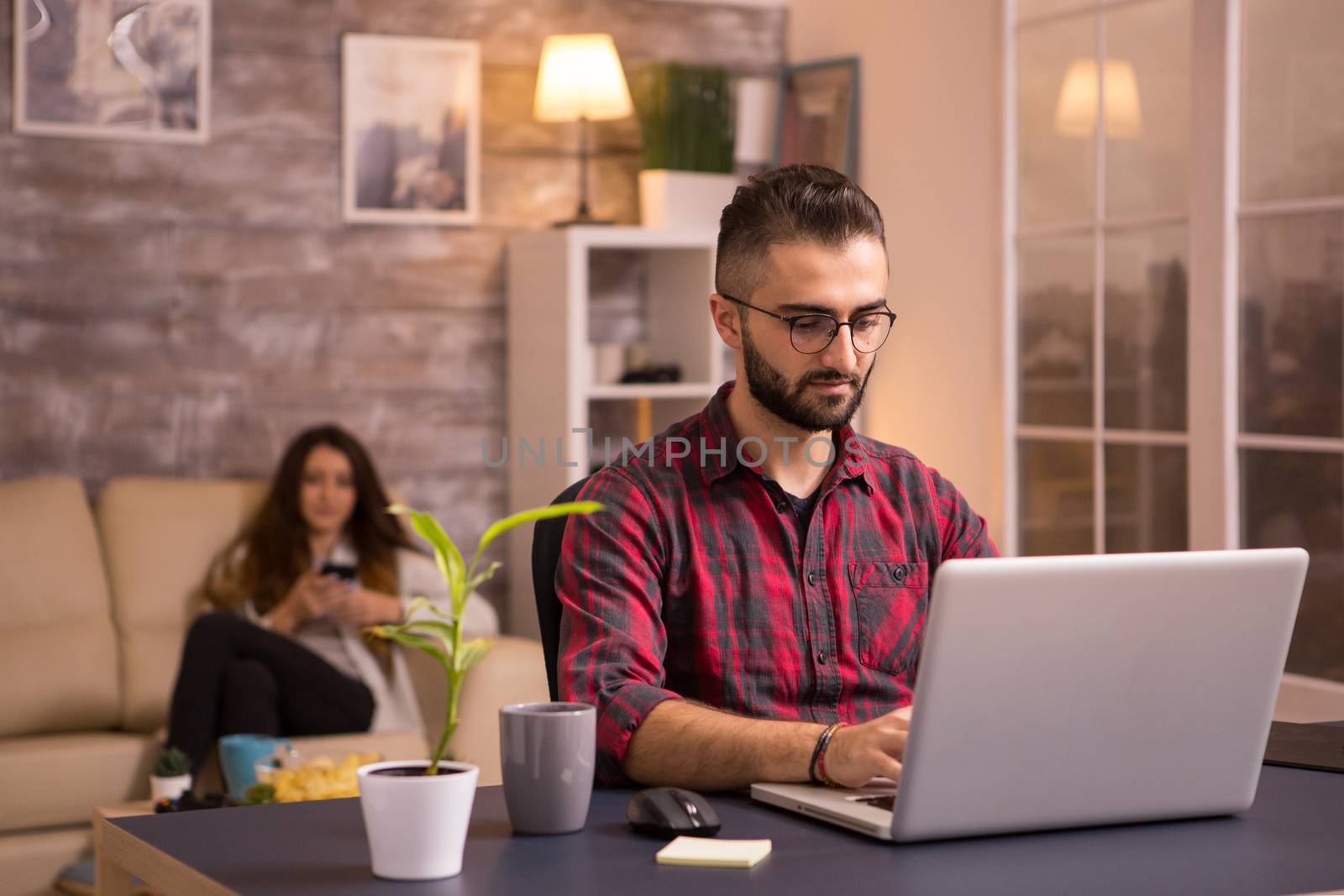 Bearded freelancer working on laptop in living room. Girlfriend relaxing on sofa in the background using her phone. Chips.