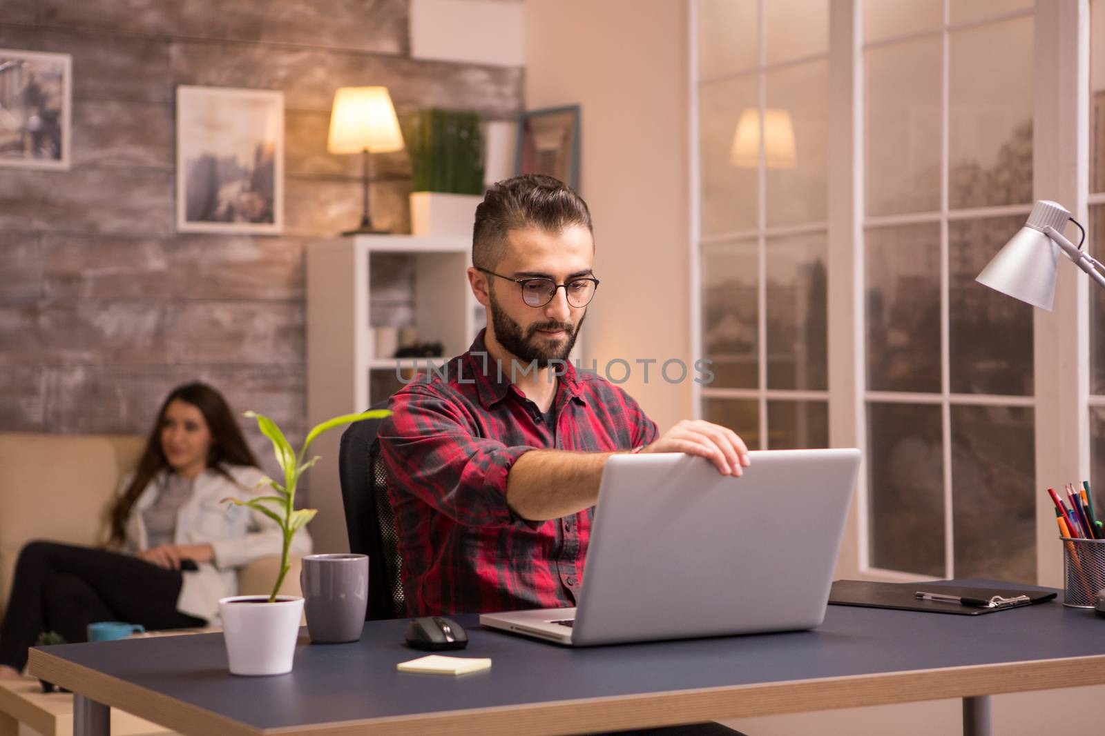 Bearded freelancer opening his laptop to start working on living room. Girlfriend relaxing on sofa watching tv in the background.