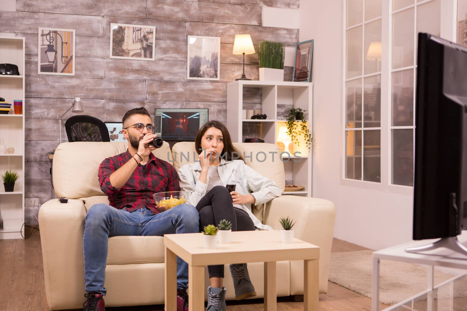 Beautiful young couple drinking soda and eating chips while watching a movie on tv sitting on couch.