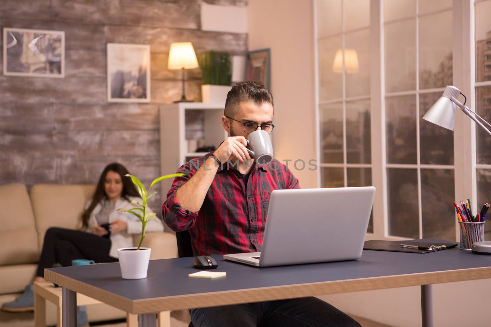 Handsome young man enjoying a cup of coffee while working on laptop from home. Girlfriend relaxing on sofa browsing on her phone.