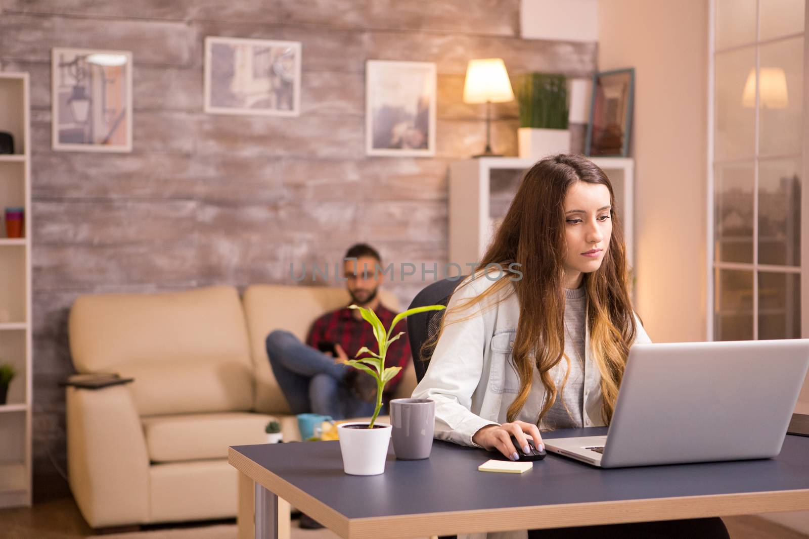 Young female working on laptop in living room and boyfriend relaxing on couch browsing on phone.