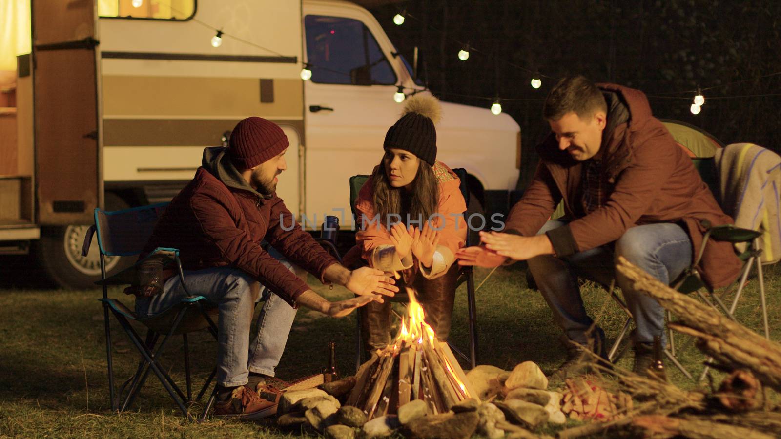 Close friend sitting on camping chairs around camp fire warming their hands. Retro camper. Light bulbs in the background.