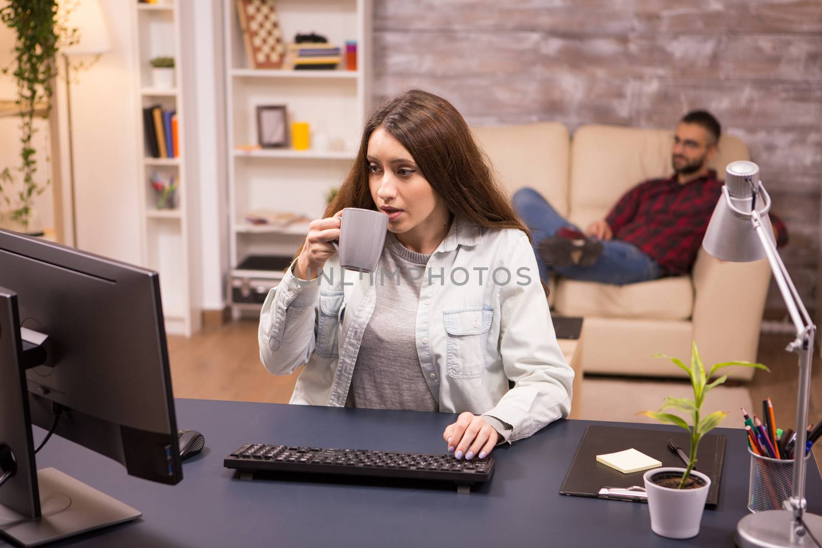 Beautiful freelancer drinking coffee while working on computer from home. Boyfriend relaxing on sofa in the background.