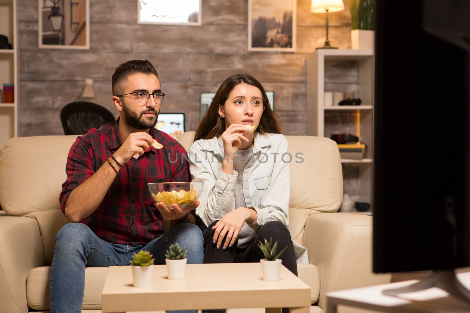 Beautiful young couple looking worried at tv while watching a movie. Couple eating chips sitting on sofa.