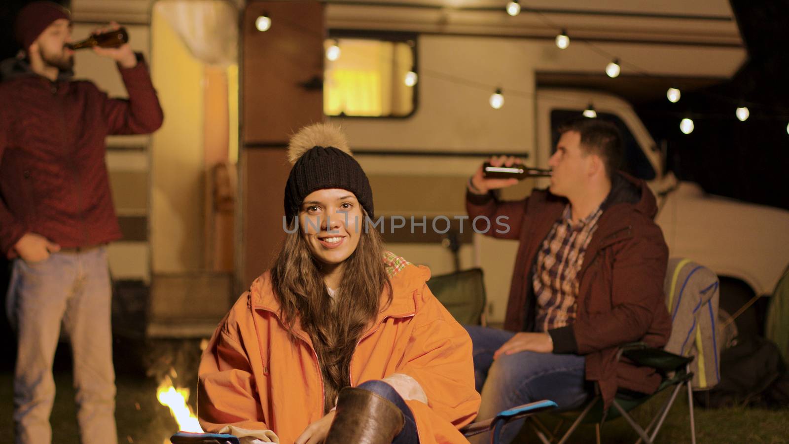 Girl sitting on a camping chair in a cold night of autumn. Friends clinking beer bottles in the background.