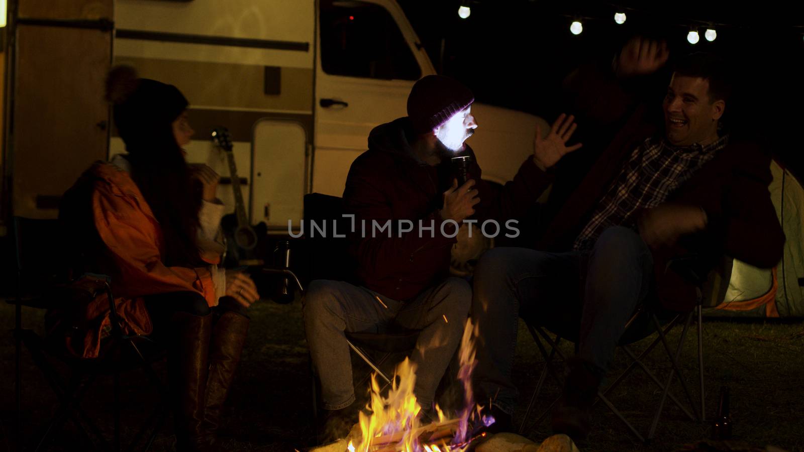 Bearded man telling a scary story to his friends around camp fire in a cold night of autumn. Retro camper van. Light bulbs