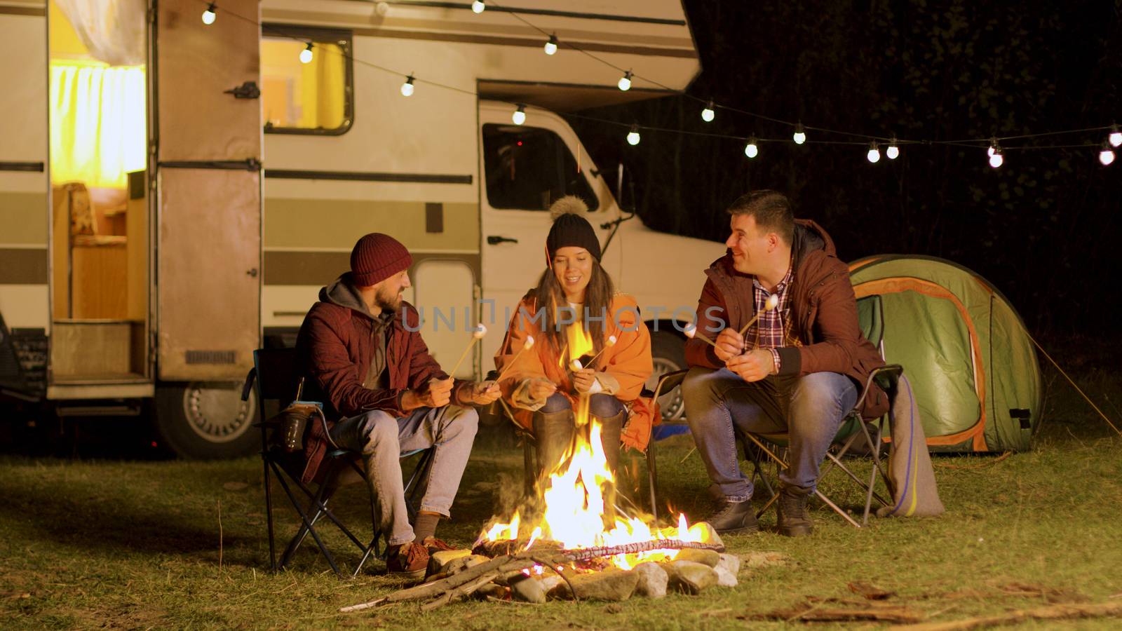 Group of happy friends around burning camping fire in the woods roasting marshmallows. Retro camper van.