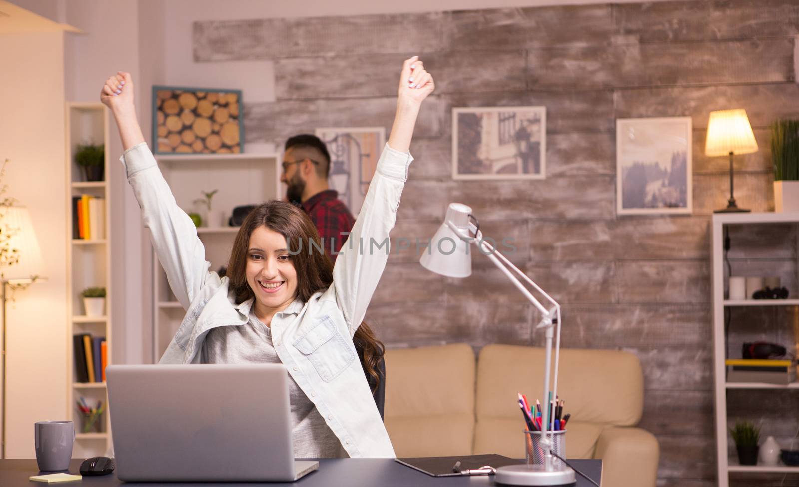 Excited female freelancer with hands raised after reading a good news on laptop. Boyfriend talking on the phone in the background.