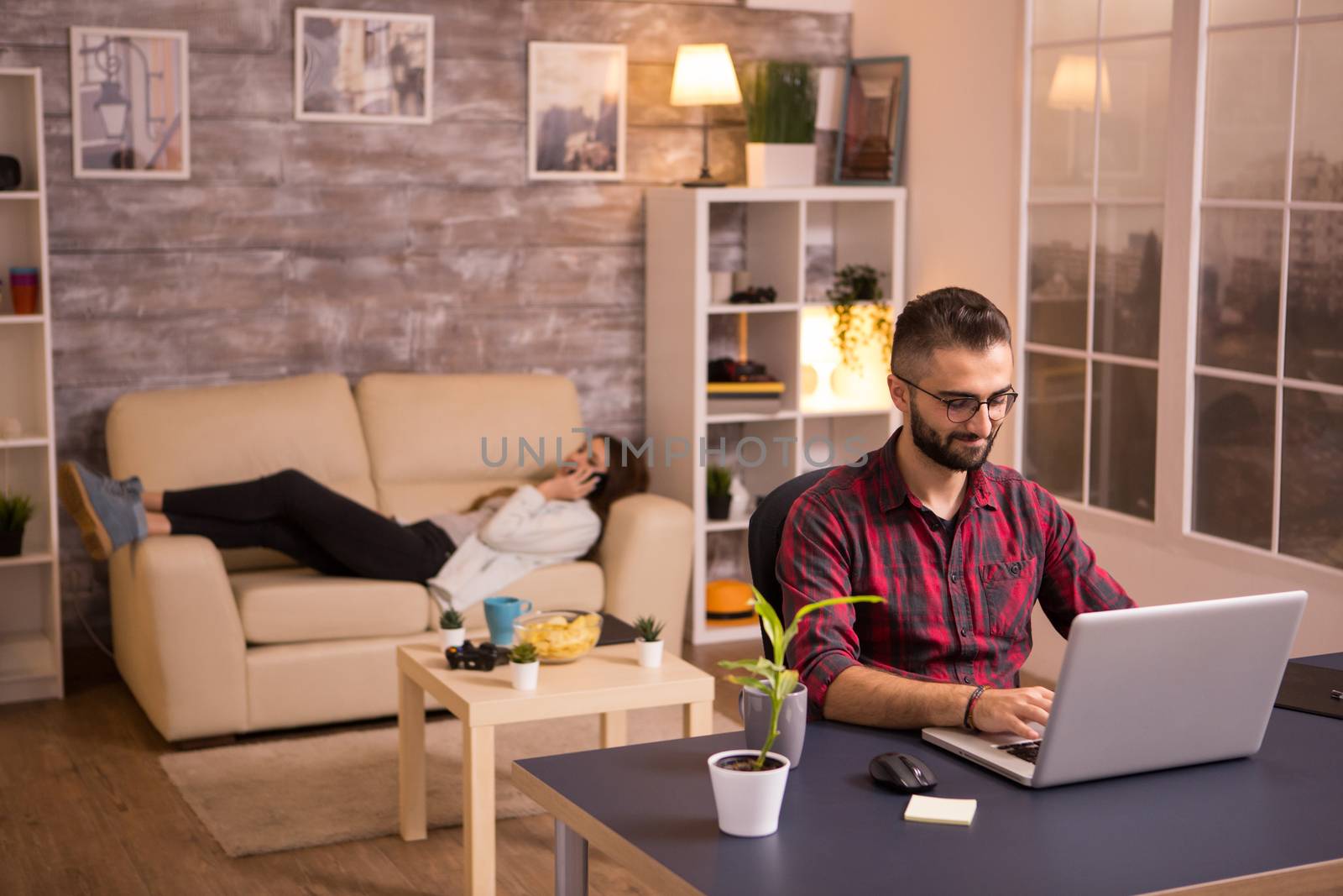 Male entrepreneur working on laptop in living room with girlfriend relaxing on sofa in the background. Chips on table.