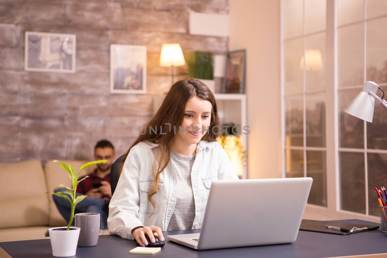 Beautiful young woman working on laptop and smiling. Woman working from home. Boyfriend relaxing on sofa in the background.