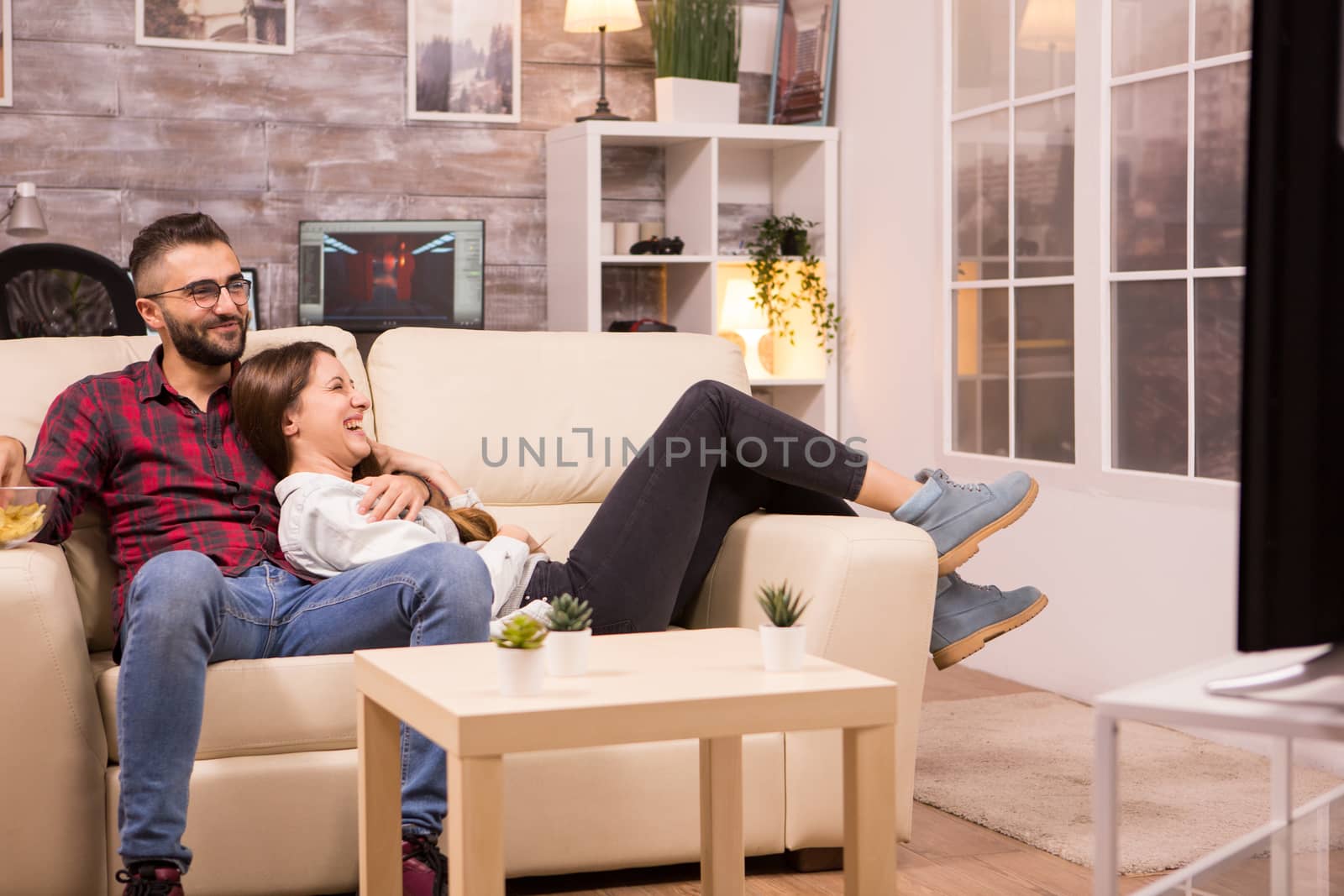 Couple laughing while watching a movie on tv and sitting on couch by DCStudio