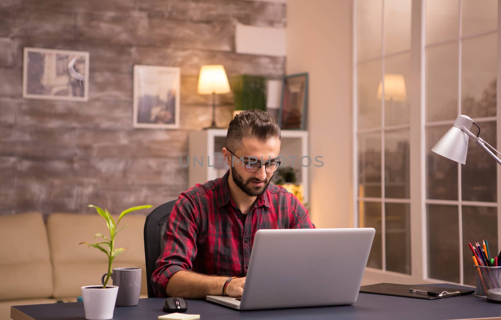 Bearded freelancer smiling while working on laptop from home. Girlfriend working in the background talking on her phone.