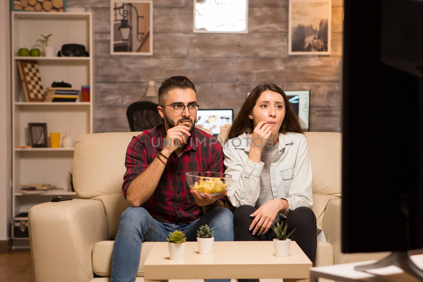 Young couple looking concentrated on tv while watching a movie and eating chips . Couple sitting on sofa.