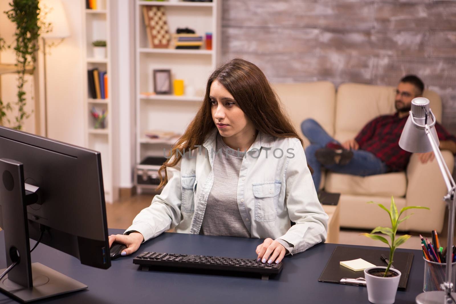 Female freelancer working on computer from home. Boyfriend relaxing on sofa in the background.