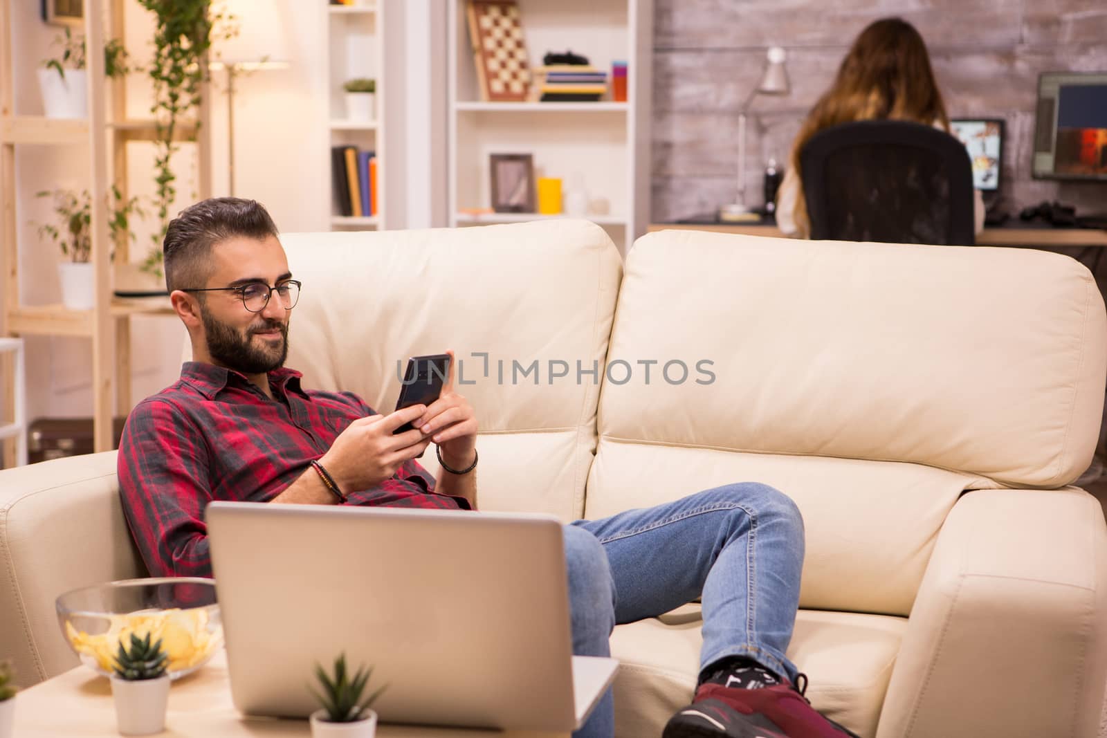 Bearded freelancer sitting on couch and browsing on his phone. Girlfriend in the background.