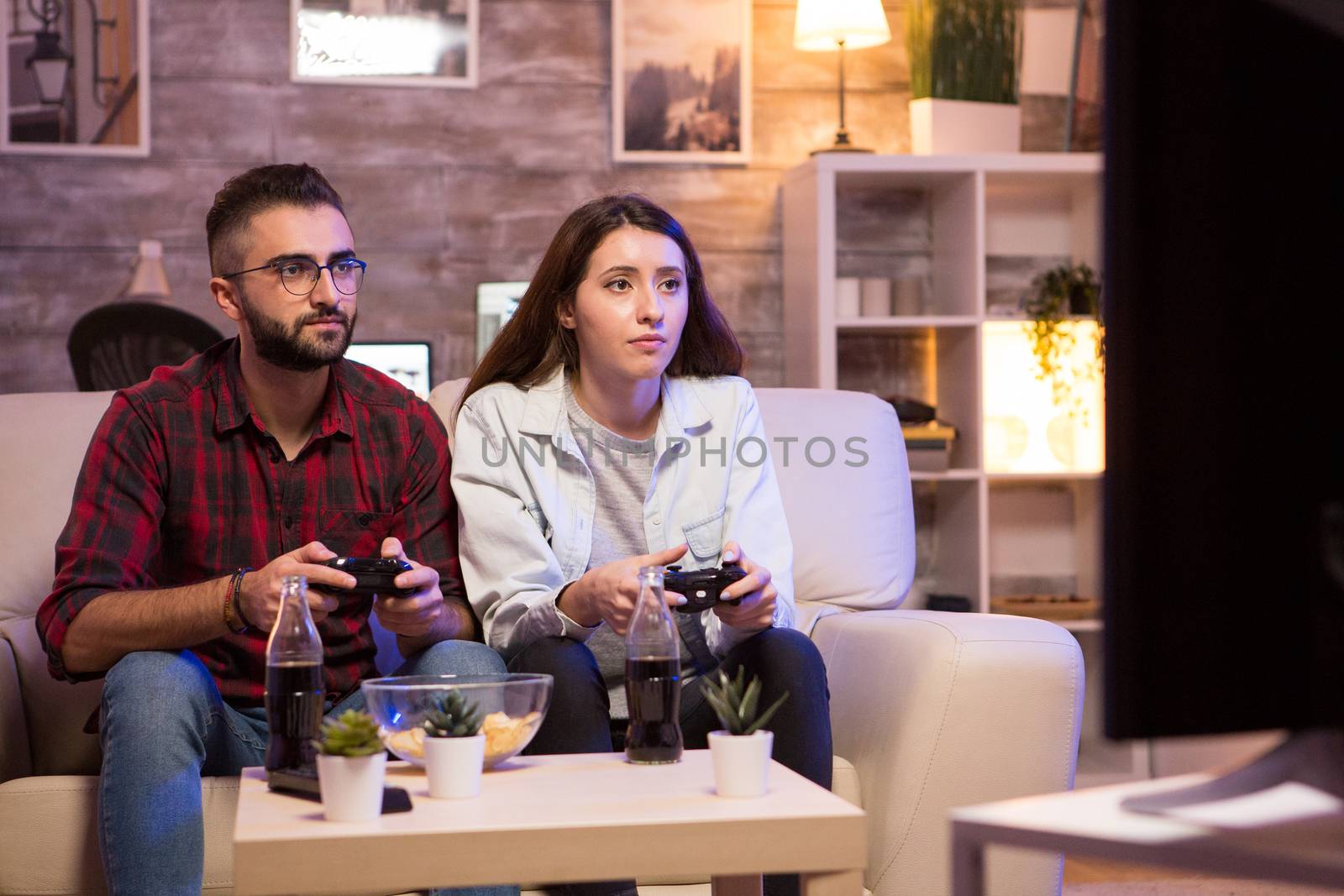 Beautiful young couple having fun playing video games with controllers at night. Couple sitting on sofa.