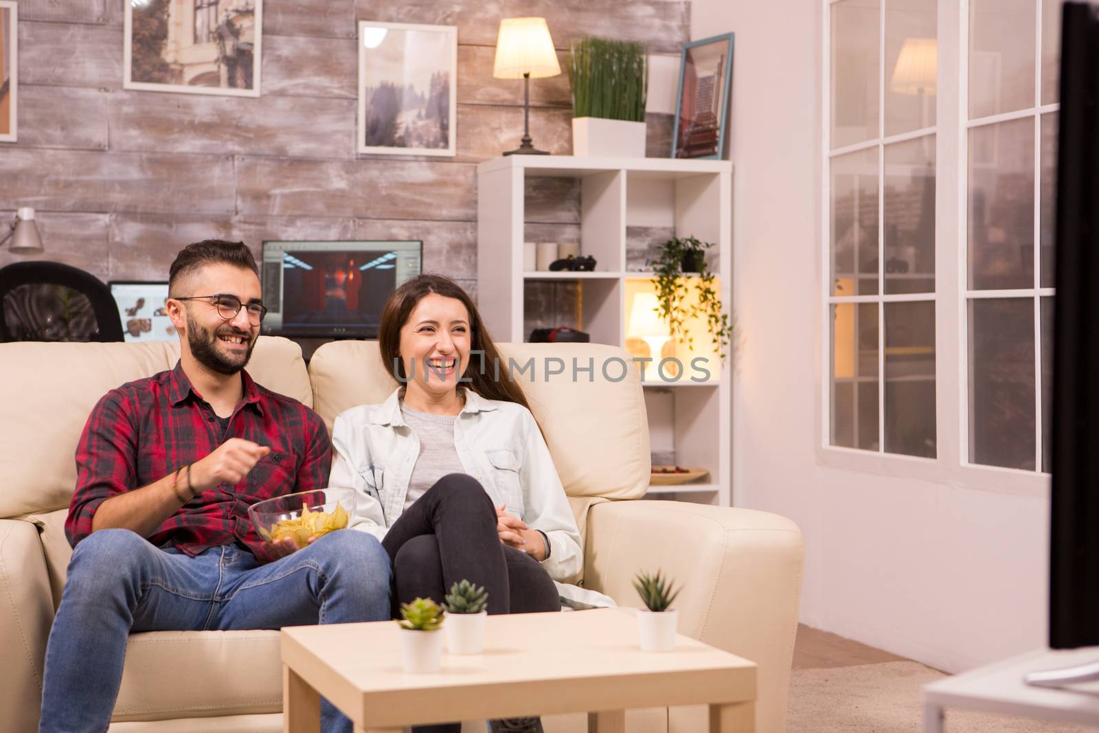 Couple laughing so hard while watching a movie on tv and eating chips.