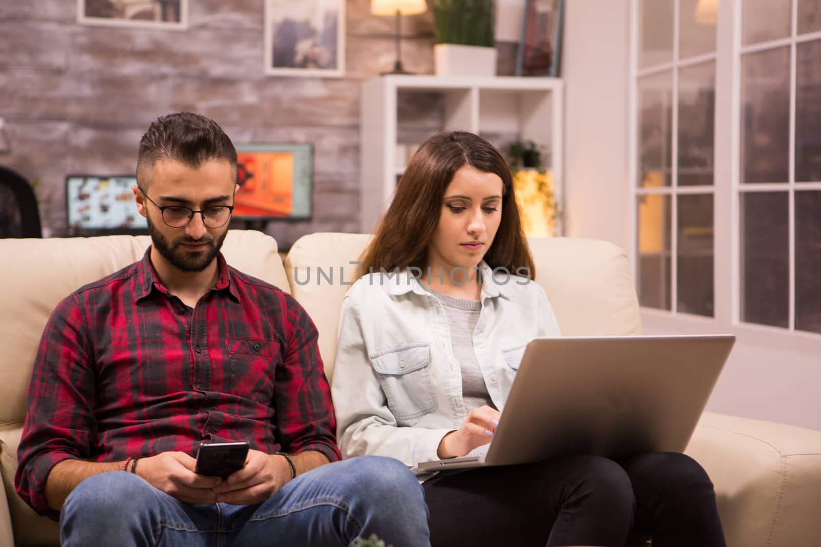 Young couple sitting on sofa and doing online shopping using their laptop and phone. Girlfriend browsing on laptop.