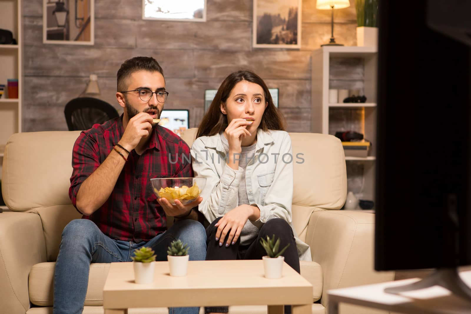 Couple eating chips and looking shocked at tv while watching a movie by DCStudio