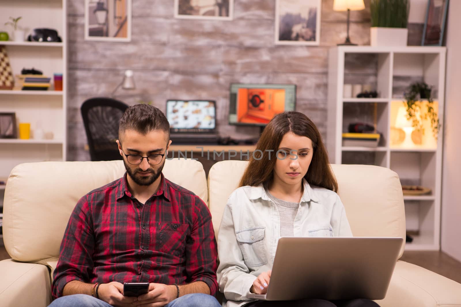 Caucasian beautiful couple with serious faces sitting on sofa. Couple using laptop and phone while sitting on sofa.