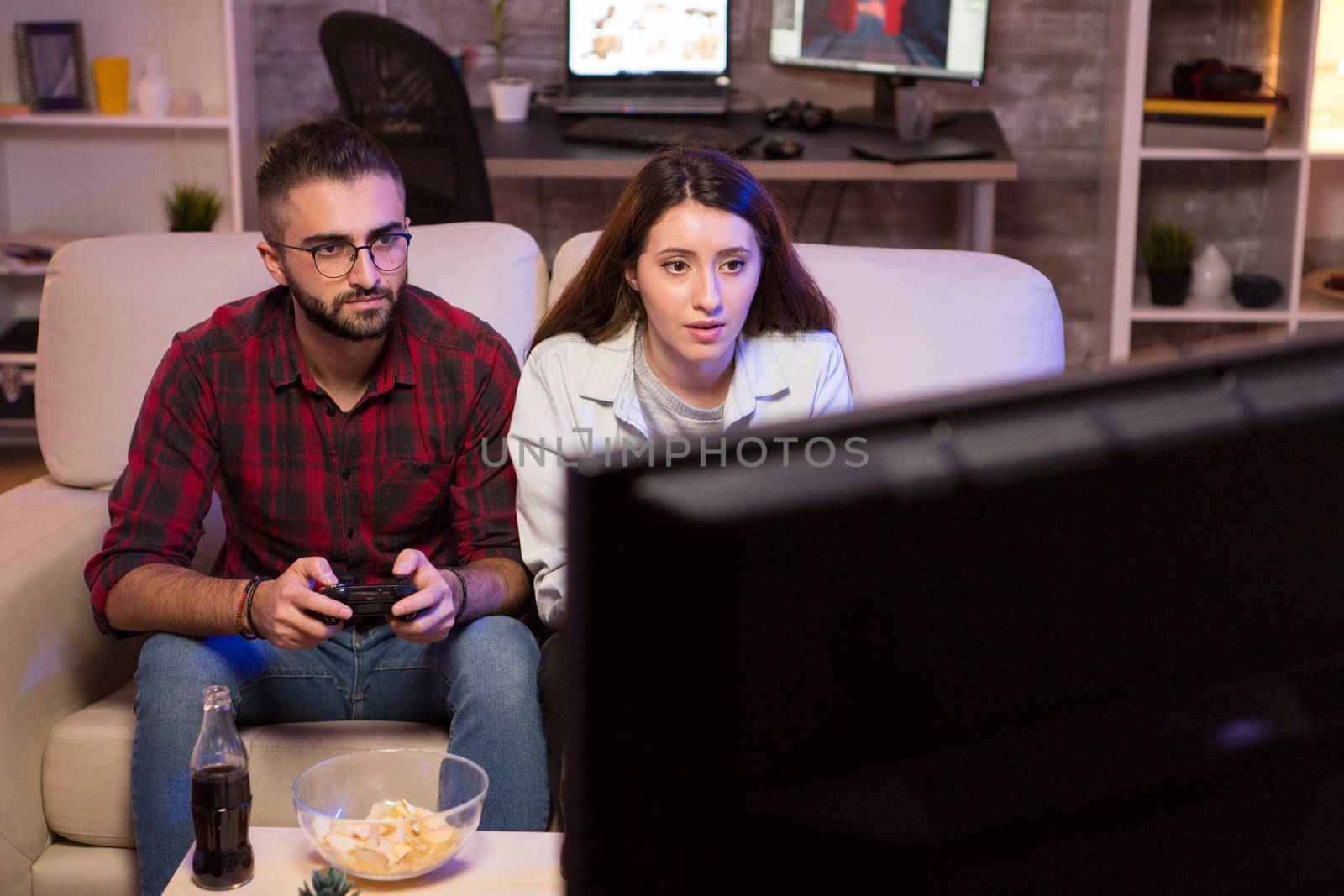 Joyful young couple playing video games on television using controllers at night. Couple sitting on couch