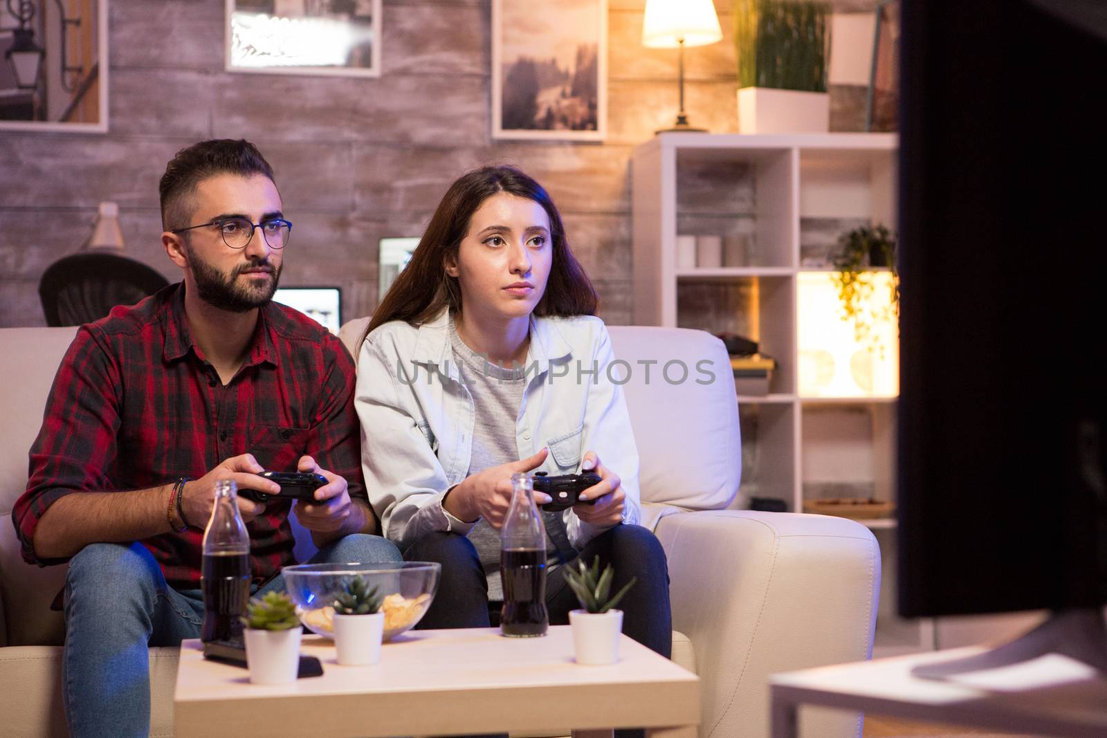 Beautiful young couple having fun playing video games with controllers at night. Couple sitting on sofa.