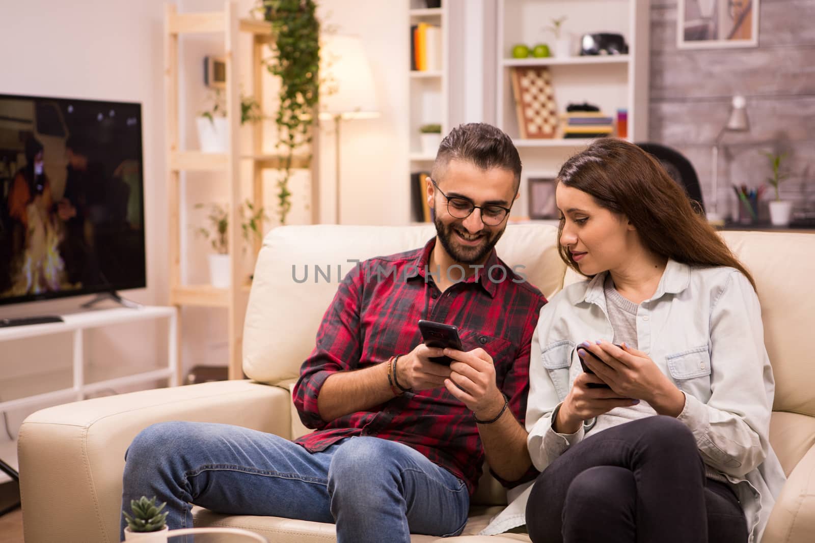 Caucasian man showing something to his girlfriend on the phone while relaxing on sofa. Couple laughing.