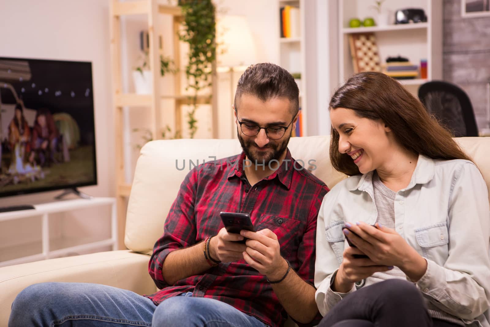 Caucasian man showing something to his girlfriend on the phone while relaxing on sofa. Couple laughing.