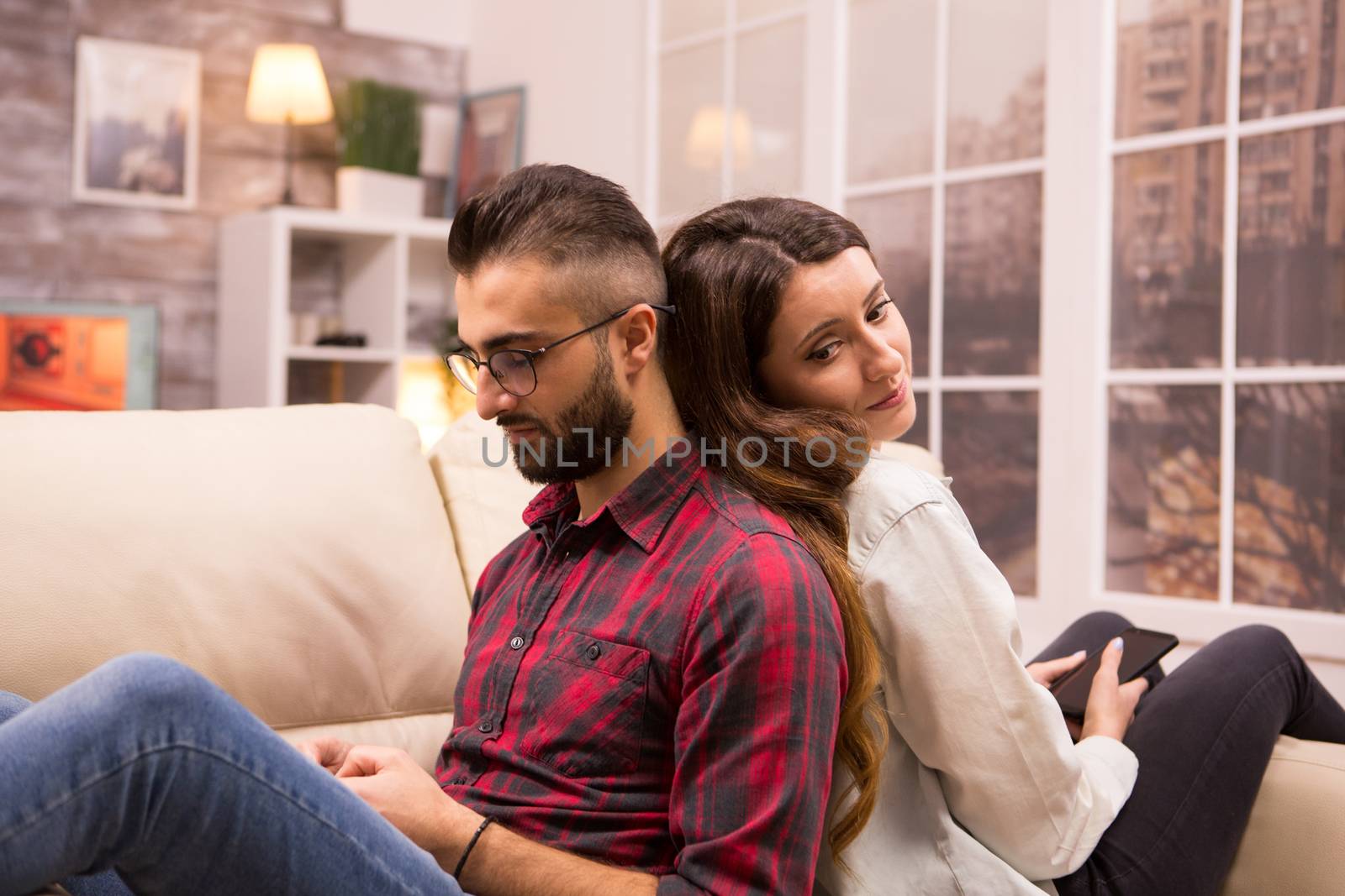Girlfriend looking at her boyfriend while sitting back to back on sofa after and disagreement. Boyfriend ignoring girlfriend.