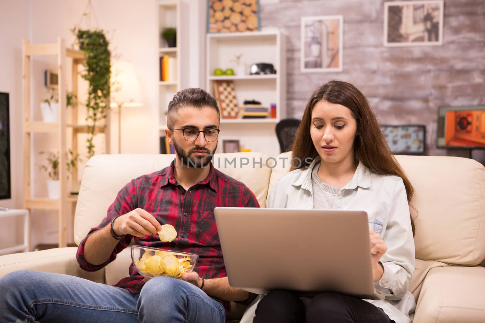 Boyfriend eating chips while his girlfriend is browsing on laptop. Couple sitting on couch.