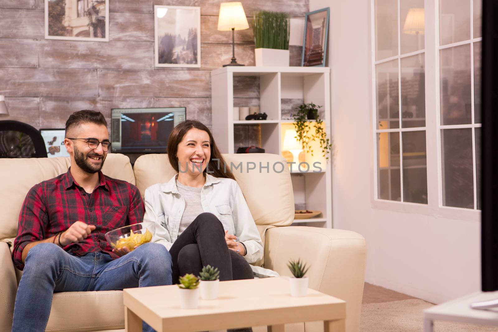 Couple laughing so hard while watching a movie on tv and eating chips.