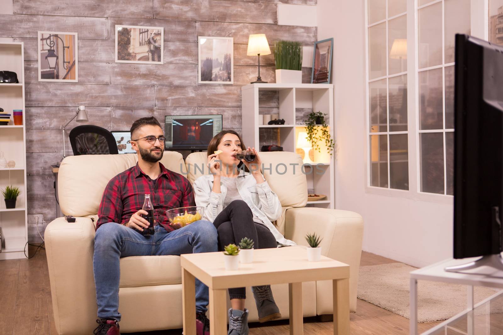 Cheerful young couple sitting on sofa and watching a movie on tv while eating chips and drinking soda.