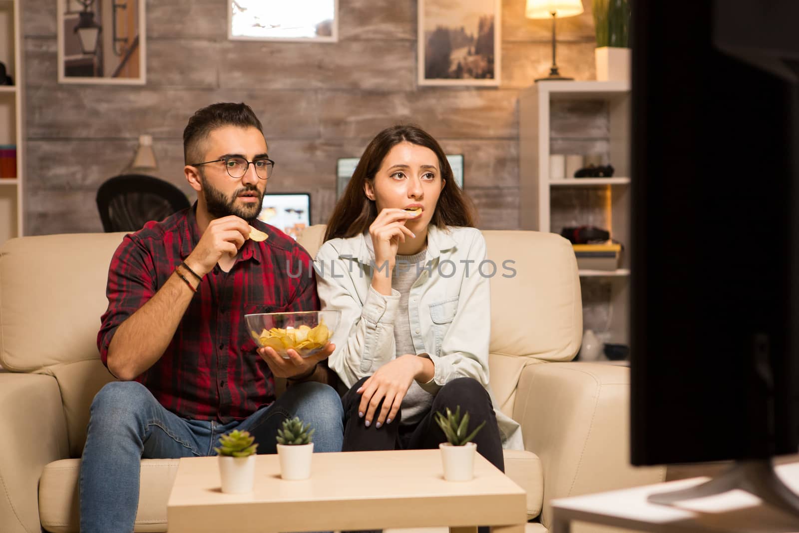 Couple eating chips and looking shocked at tv while watching a movie by DCStudio