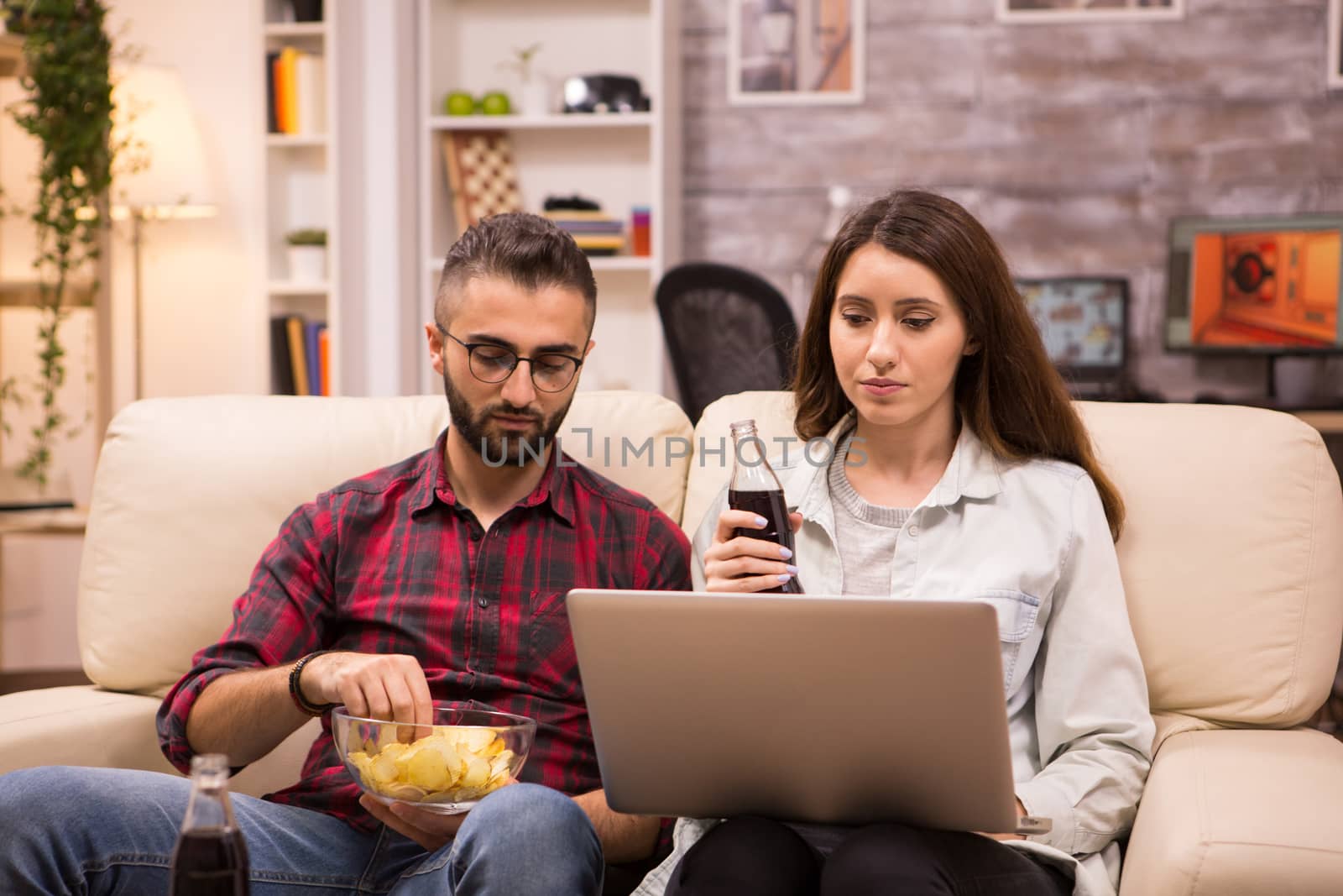 Happy young couple drinking soda and eating chips while watching a movie on laptop. Couple sitting on sofa.