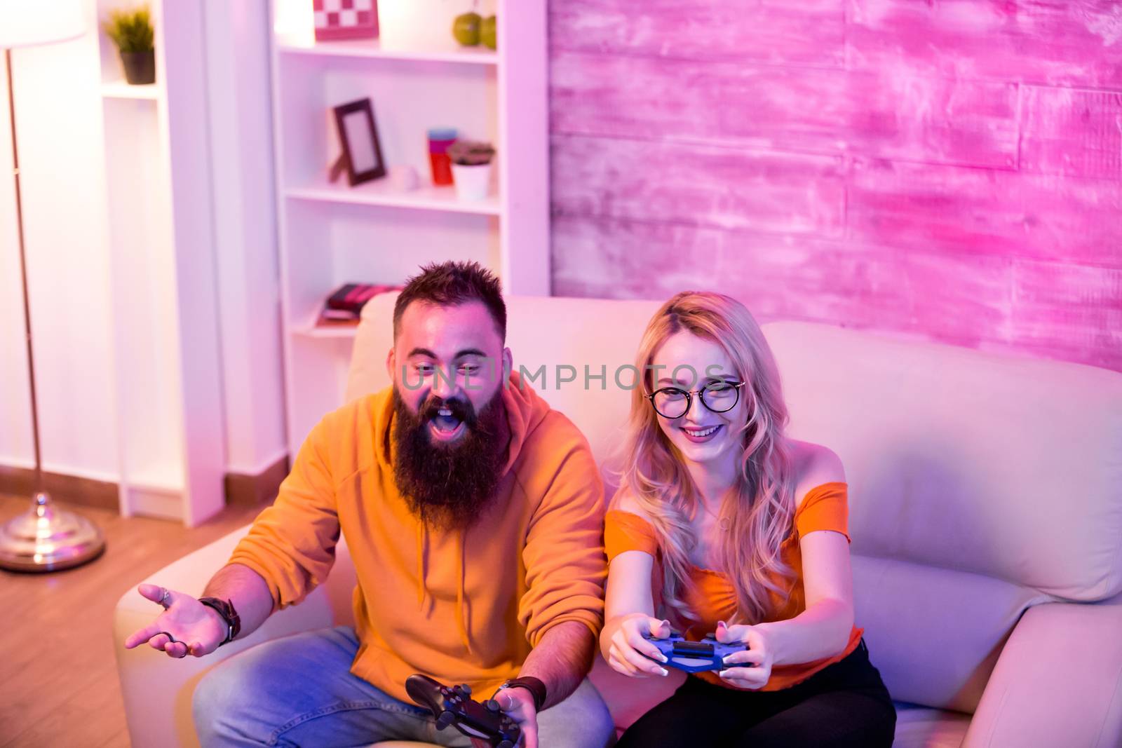 Boyfriend can't believe his girlfriend is winning at online video games. Couple sitting on sofa. Neon light in the room.