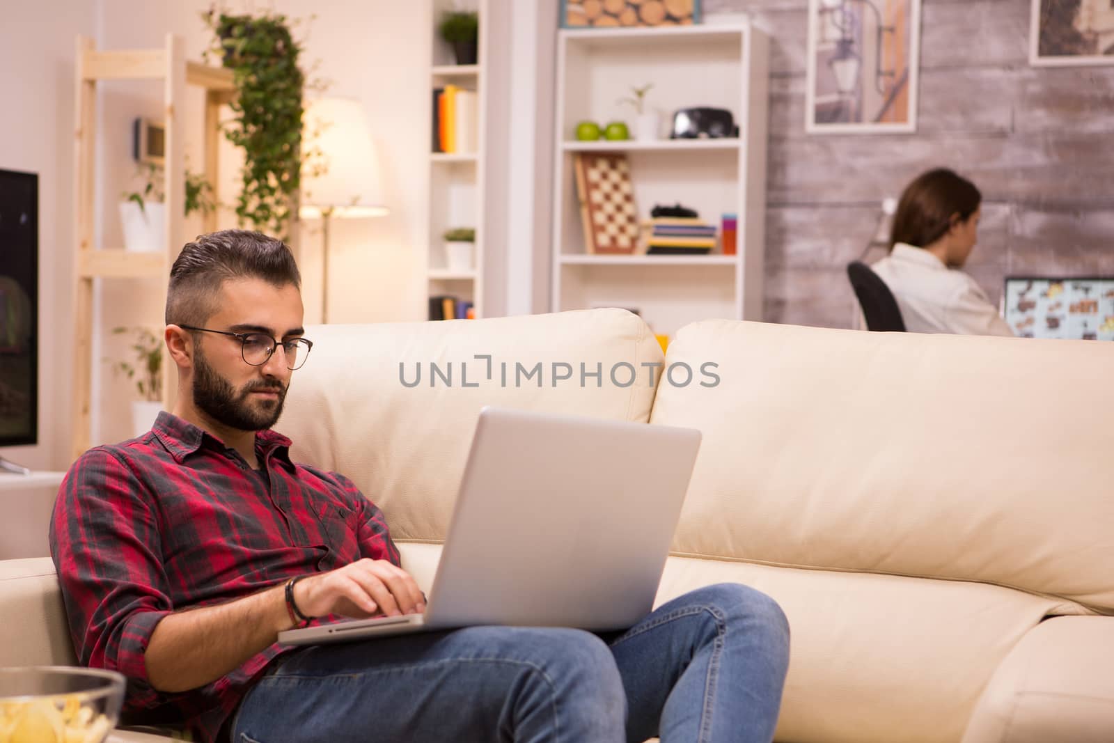 Young entrepreneur working on laptop sitting on sofa. Girlfriend in the background.