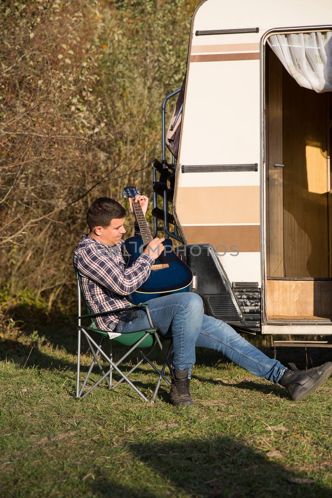 Carefree man relaxing playing on his guitar in the mountains in front of his retro camper van.