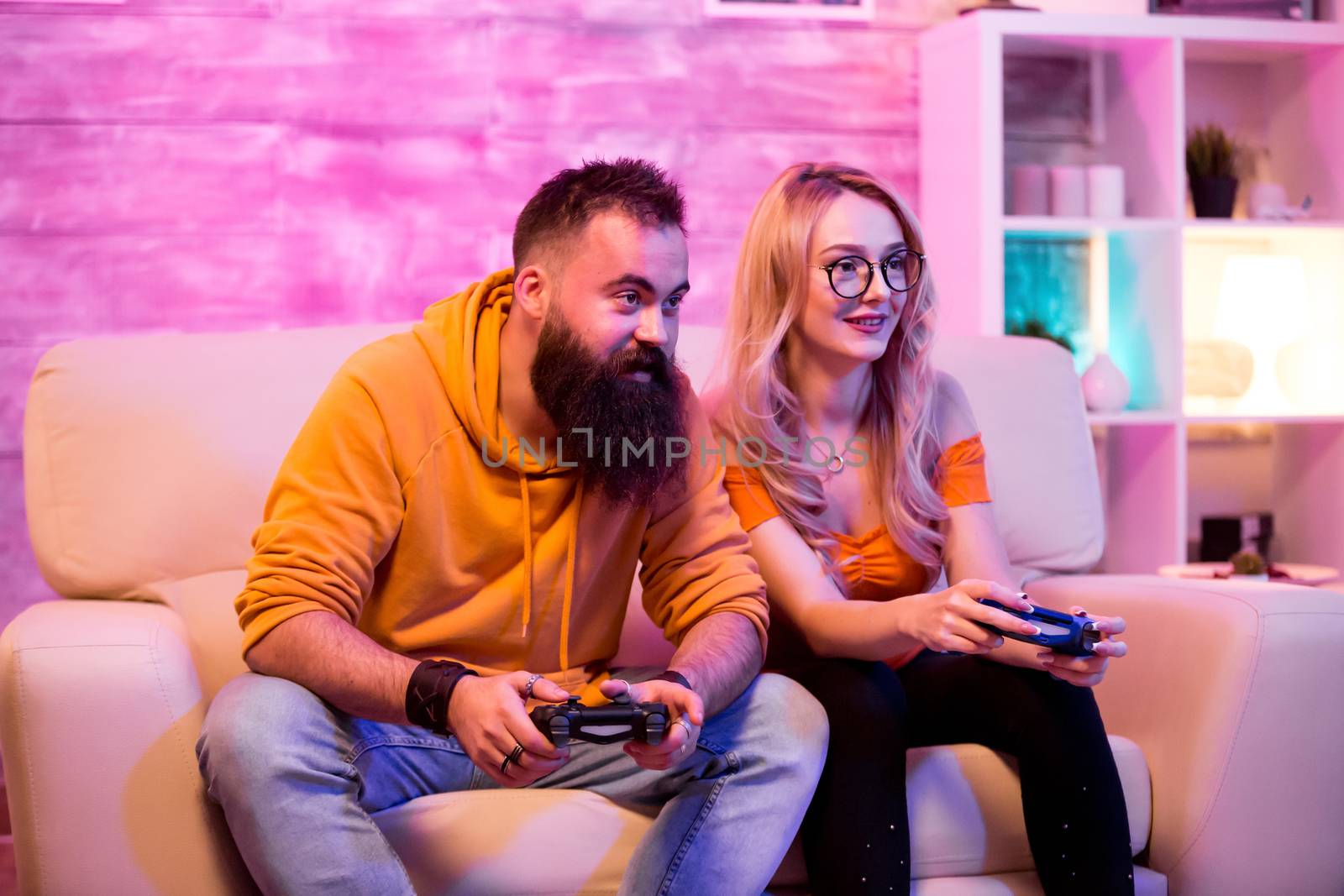 Young man and woman using wireless controller to play online games sitting on sofa. Room with neon light.