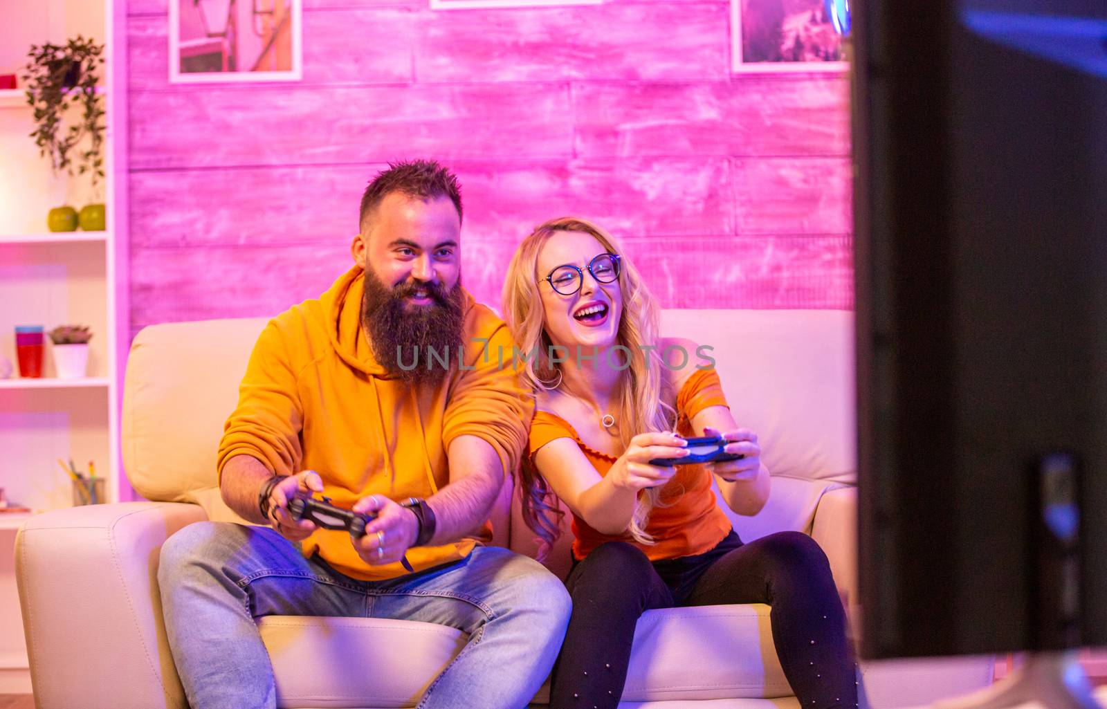 Beautiful blonde girl smiling while playing video games with her boyfriend using wireless controllers on a big screen TV