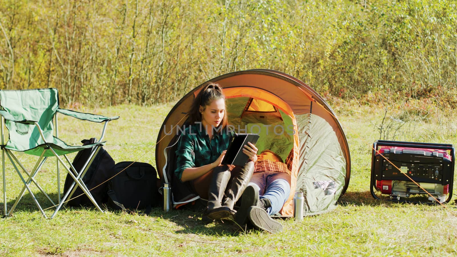 Beautiful young girlfriend using her tablet in front of camping tent while her boyfriend's relaxing inside the tent.