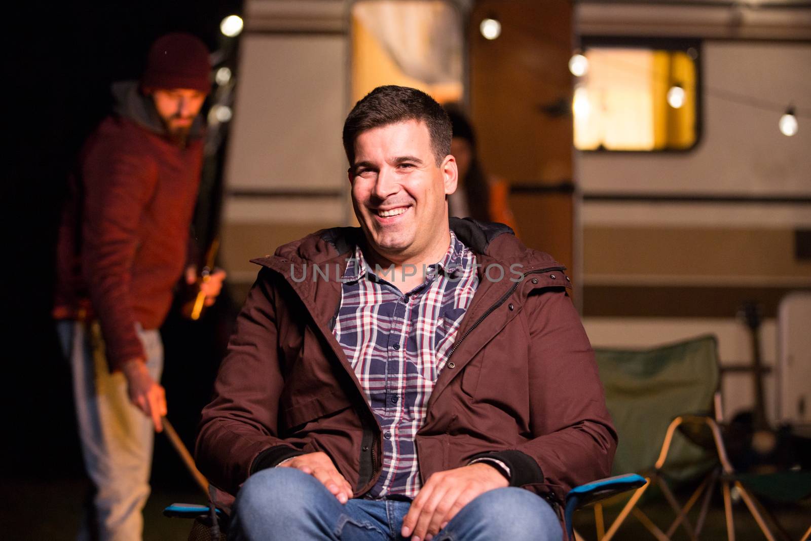 Portrait of young man smiling and sitting on camping chiar in a campsite. Retro camper van in the background.