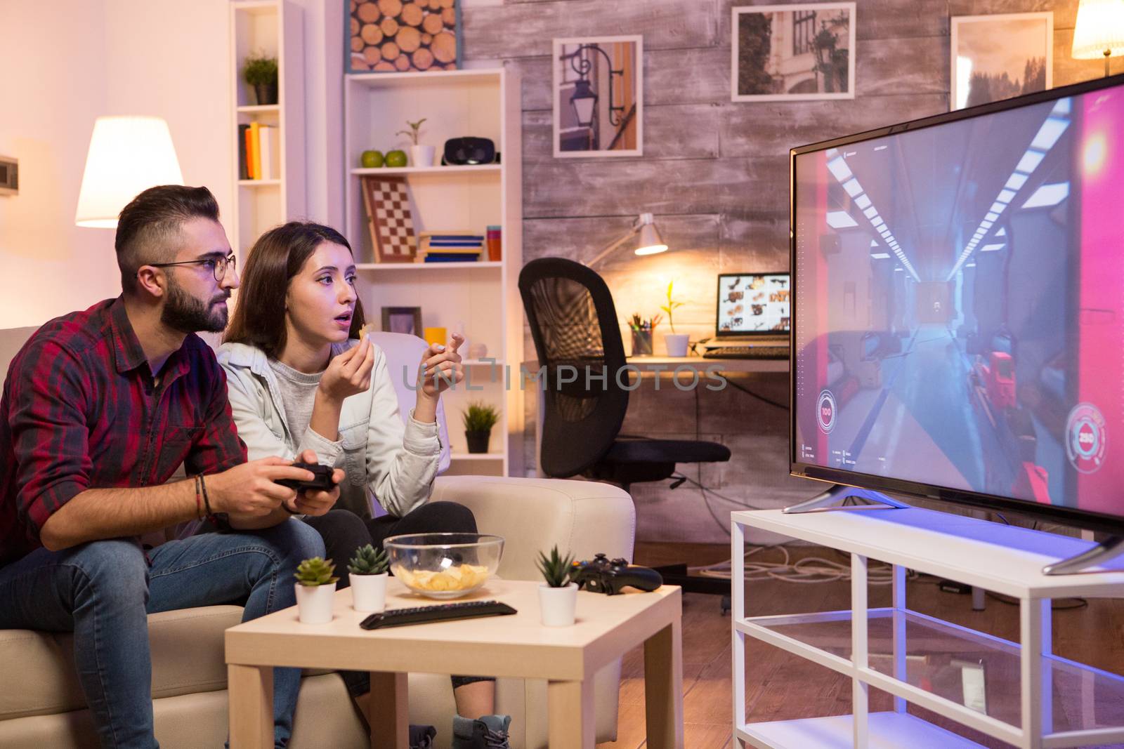 Boyfriend playing video games on television using controller while girlfriend is eating chips. Couple sitting on sofa.