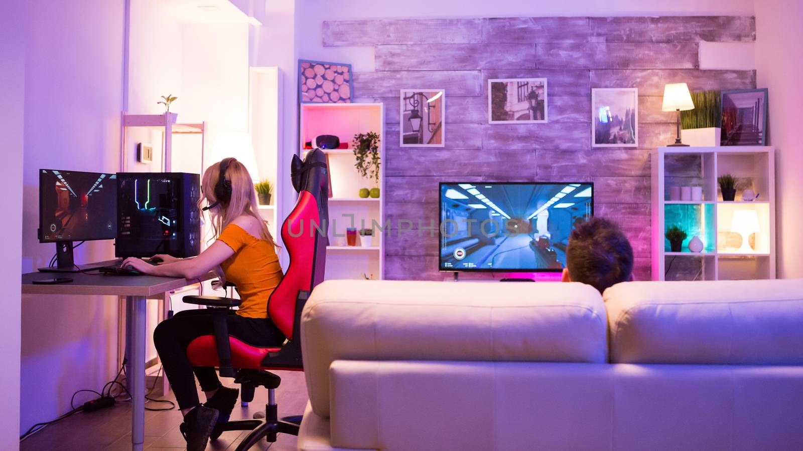 Girl with headphones sitting on gaming chair and playing shooter games. Male sitting on sofa.