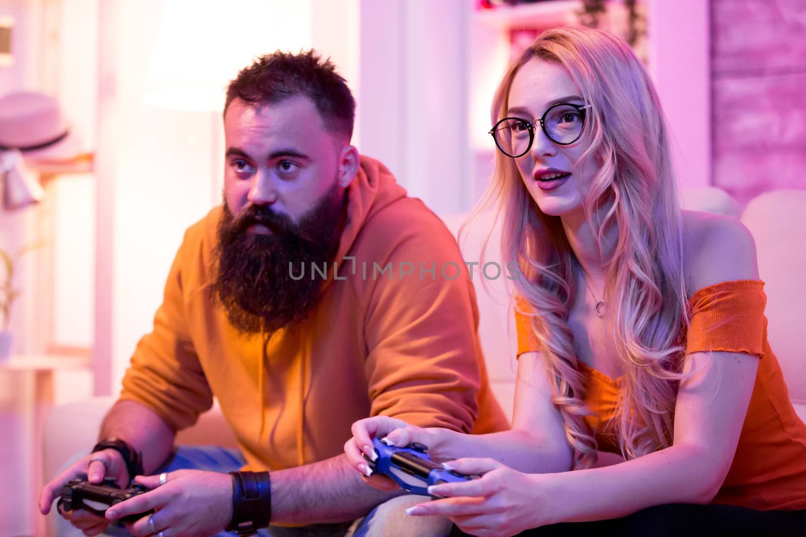 Couple sitting on sofa and playing online video games using wireless controler. Room with neon light.