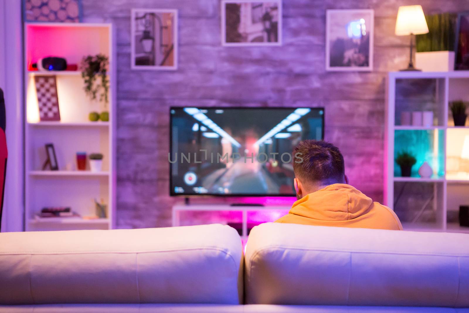 Back view of young man playing video games on television sitting on sofa.